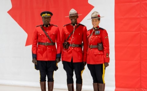 Devant une murale du drapeau canadien, deux policiers et une policière vêtus de tuniques rouges sourient à l'appareil photo.