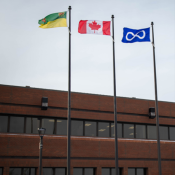 Trois drapeaux—Saskatchewan, Canada et Métis—flottant sur des poteaux devant un immeuble en briques, sous un ciel couvert.