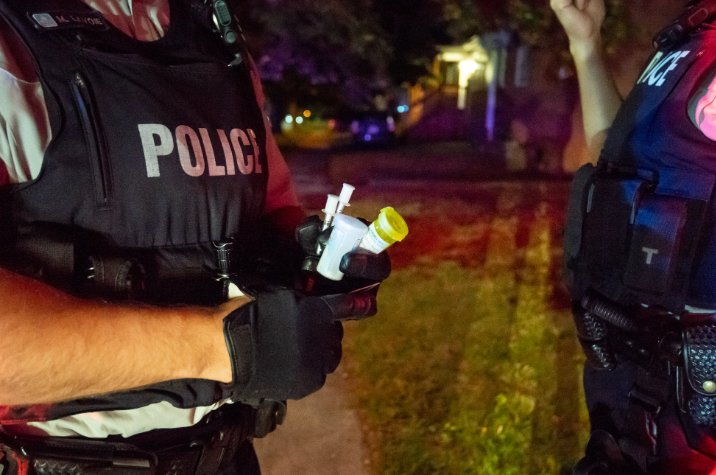 Gros plan de policiers de la GRC la nuit lors d'un contrôle routier; un des policiers tient une bouteille de médicaments. Les gyrophares projettent des lueurs rouges et bleues en arrière-plan. 