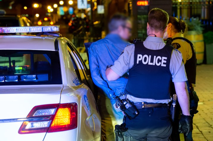 Des policiers de la GRC sont debout à côté d'un véhicule de police identifié pendant un contrôle routier, la nuit, et un individu se trouve près de l'arrière du véhicule. Des lampadaires et un environnement urbain sont visibles en arrière-plan.