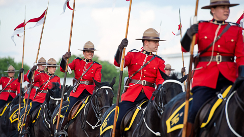 Plusieurs agents de la GRC vêtus de la tunique rouge transportent des drapeaux rouges et blancs à dos de chevaux noirs.