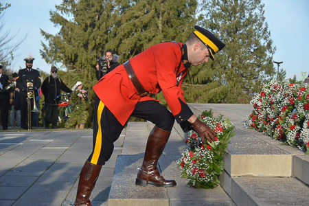 Un agent de la GRC en tunique rouge se penche pour déposer une couronne au pied d’un monument. On voit également d’autres personnes et d’autres couronnes.