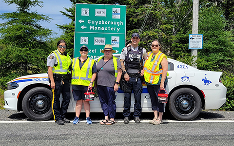 Cinq personnes, dont deux agents de police, se tiennent devant une voiture de police et des panneaux de signalisation routière.