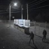Two individuals, dressed all in dark clothing with hoods over their heads, walk into a fenced compound at night. A power substation is visible in the background.