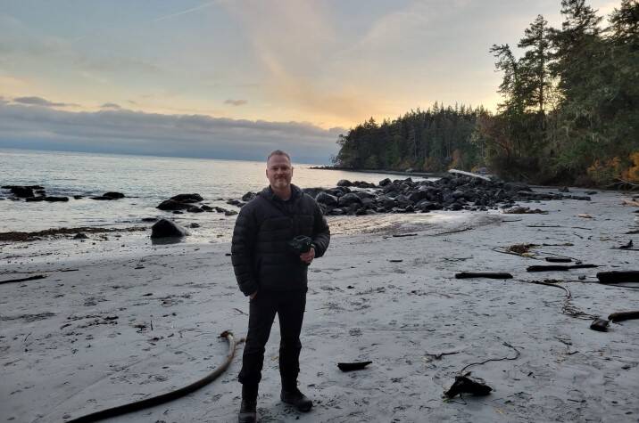 Man dressed in light outwear stands on a rocky beach with trees and a sunrise in the distance.
