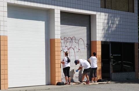 Three people work to remove graffiti from a corrugated metal sliding door at a commercial building.